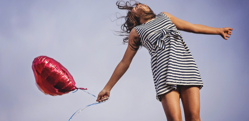 Eine Frau im Sommerkleid springt nach oben, hält einen Herz-Luftballon in der Hand.