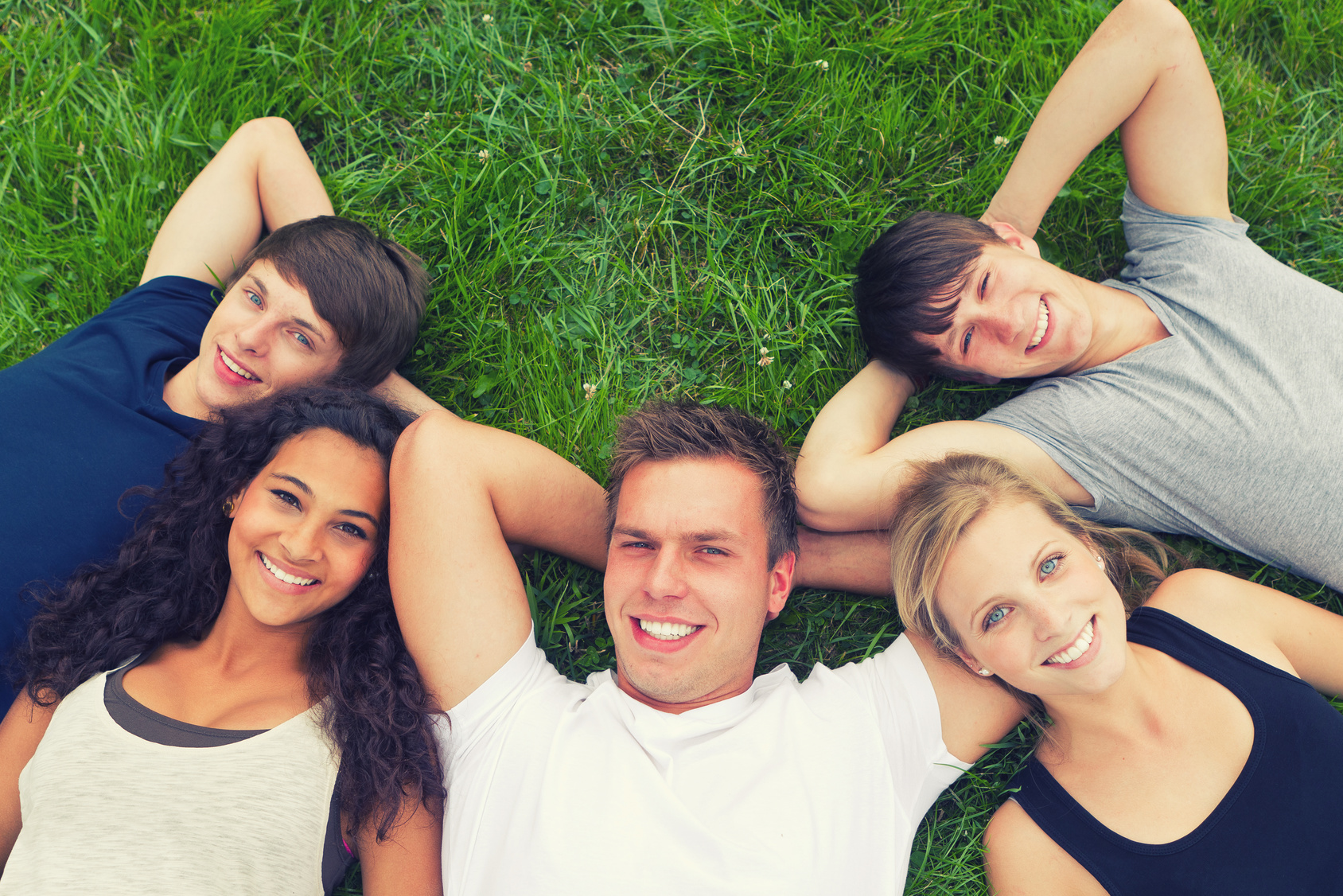 Man sieht den Oberkörper von 5 jungen Menschen, die im Gras liegen und nach oben lächeln. 